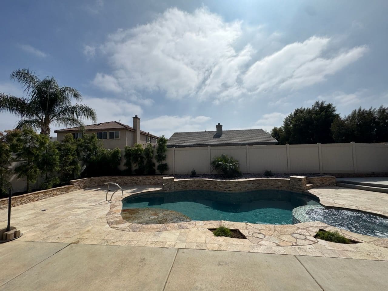 Backyard pool with stone deck and lush greenery under a blue sky with clouds.